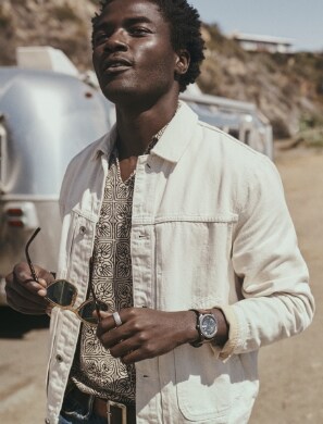 Man in white denim jacket wearing a mens Fossil watch and holding Fossil sunglasses in front of an Airstream trailer.