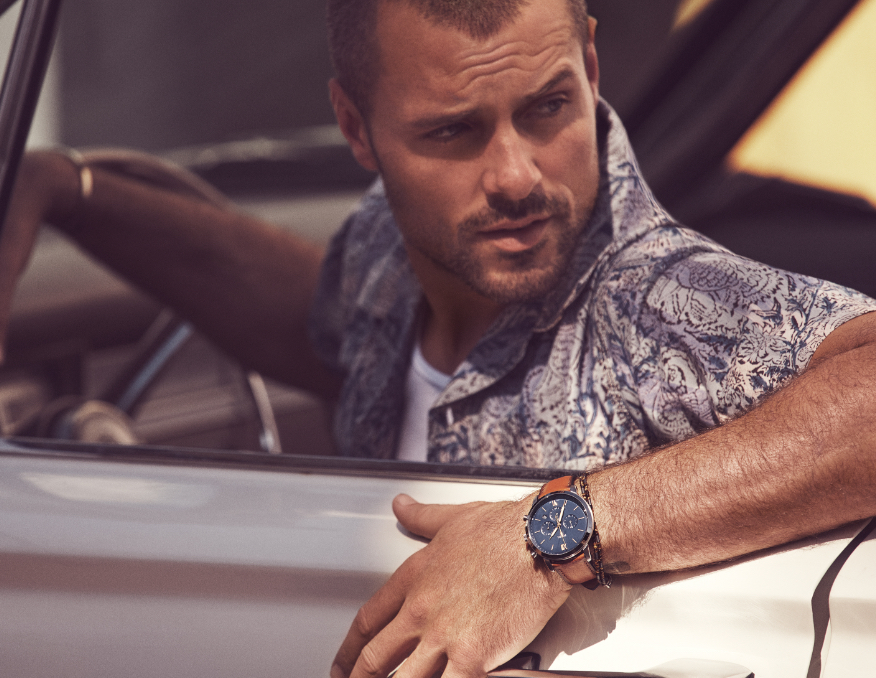 Stylish man and woman driving car wearing gold and silver-tone ladies watch and brown leather men's watch with a blue dial.
